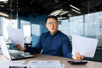 Fototapeta na wymiar Problems at work. Worried young Asian man, businessman, freelancer. He sits in the office at a table with a laptop, holds documents in his hands. Spreads his hands, looks at the camera confused.