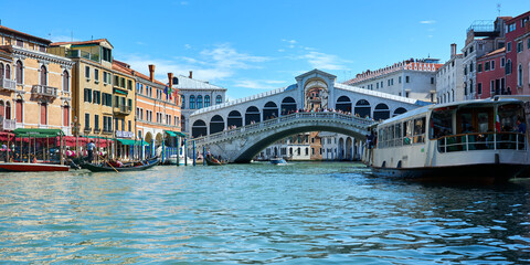 Beautiful panoramic cityscape in Venice, Italy, with picturesque old buildings surrounding the Rialto Bridge on the Grand Canal, accompanied by gondolas and boats under vivid blue sky.