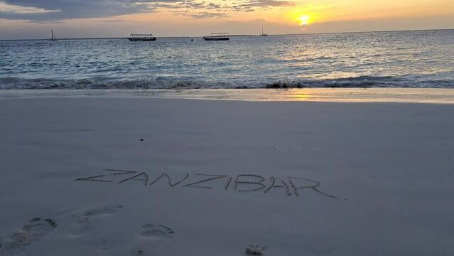 Sign ZANZIBAR on the beach with the ocean, some boats sailing at sunset
