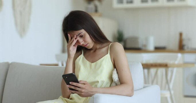 Upset young woman sits on sofa with cell phone, receive sms with bad news, read unpleasant message from boyfriend, missed or waiting for important call, staring at smartphone screen feels frustrated