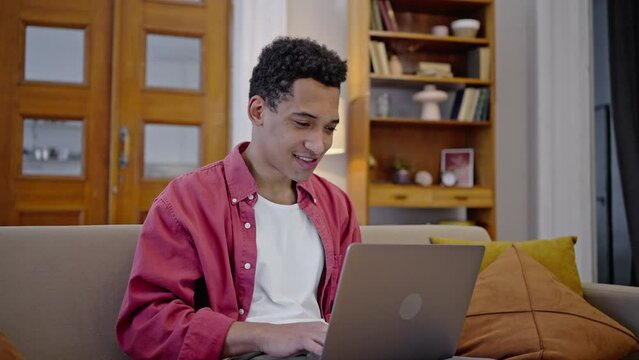 Dark-skinned guy works remotely on laptop, rejoices and performs dance movements, sitting on sofa