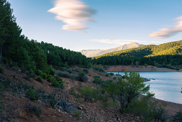 Fototapeta premium Beautiful landscape of a lake surrounded by greenery mountains and trees under a blue sky with amazing clouds at sunset, El Vado, Guadalajara, Spain