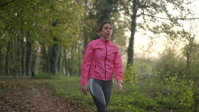 Young female wearing pink tracksuit walking and stretching her hands in an autumn forest at sunset in slow-motion