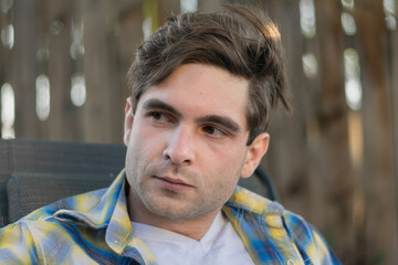 A young white man with a serious expression looks to his right outdoors in a shady backyard setting.