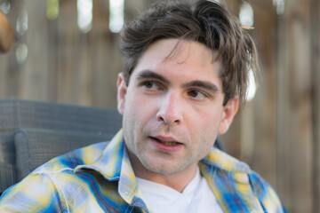 A young white man with messy hair looks to his right outdoors in a shady backyard setting.