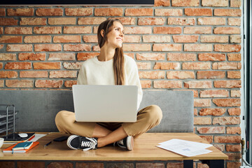 Happy young woman sitting on the desk with laptop looking out the window in contemporary loft office
