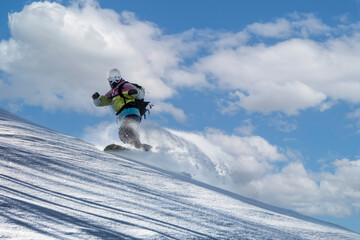 A snowboarder in a ski mask and helmet descends soft snow hills, scattering the snow