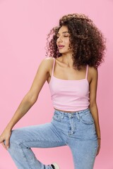 Woman with curly afro hair dancing in pink top and jeans on pink background, smile, copy place