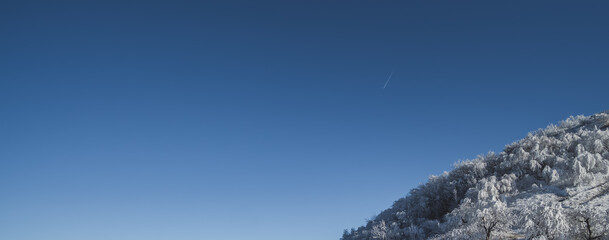 Winter panorama, the slope of Mount Beshtau with icy and snow-covered vegetation against a clear blue sky, a condensation trail from an airplane in the sky on a sunny winter day