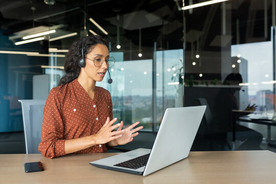 Video call online meeting with colleagues, Hispanic woman working inside modern office, businesswoman smiling and talking remotely using laptop and headset.