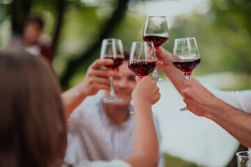 Group of happy friends toasting red wine glass while having picnic french dinner party outdoor during summer holiday vacation near the river at beautiful nature