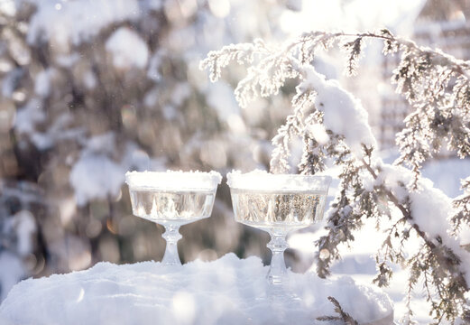 Two crystal glasses of sparkling wine or champagne cooling in snow on winter natural background with beautiful sunlight bokeh.