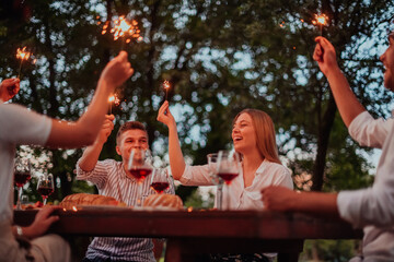 Group of happy friends celebrating holiday vacation using sprinklers and drinking red wine while having picnic french dinner party outdoor near the river on beautiful summer evening in nature