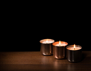 Three Brass Cup Candles, lit and placed in a row on a wooden surface with a black, in the dark background ideal for copy space.