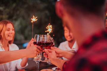 Group of happy friends celebrating holiday vacation using sprinklers and drinking red wine while having picnic french dinner party outdoor near the river on beautiful summer evening in nature