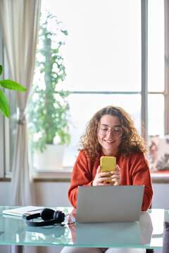 Young happy pretty woman sitting at table holding smartphone using applications on cellphone modern technology, looking at mobile phone while working or learning at home. Vertical