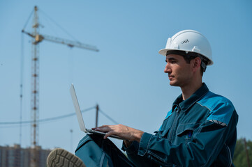 Caucasian male builder in hardhat sits on floor slabs and uses laptop at construction site. 