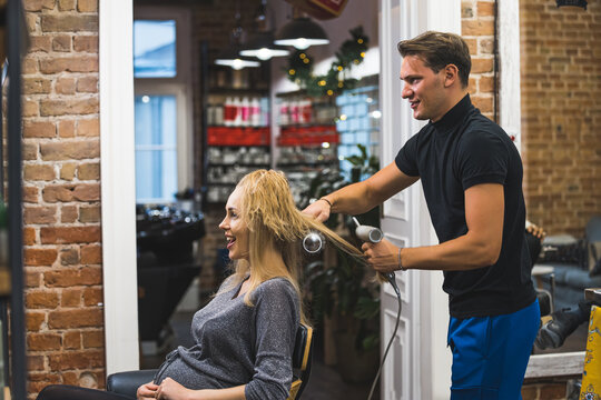 blond cheerful pregnant woman sitting on the chair and barber man drying her hair, medium shot self-care while pregnancy. High quality photo