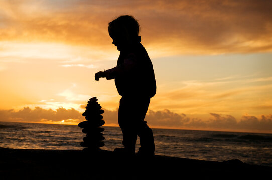 Two year old toddler stacking rocks on the beach at sunset.
