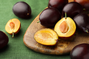Many tasty ripe plums and wooden board on green fabric, closeup