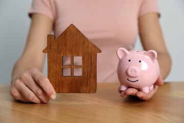 Woman holding piggy bank and house model at wooden table, closeup