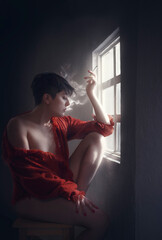 a person dressed in only an cardigan, smokes a cigarette sitting in front of a window