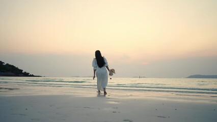 Young Asian woman in white dress walking on the beach during travel tropical island on summer holiday vacation vacation at sunset. Attractive girl relax and enjoy outdoor lifestyle walking at seaside.