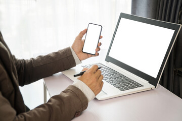 Bussinessman hand holding smartphone and pen with blank or white screen and computer laptop in office desk