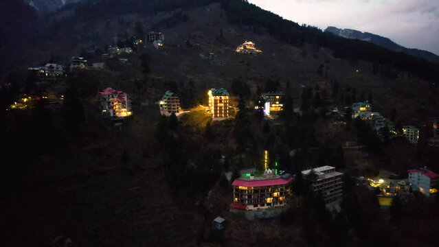 aerial drone landing shot showing lit multi floor story buildings on side of hill at night evening showing hotels shopping areas in manali, shimla himachal