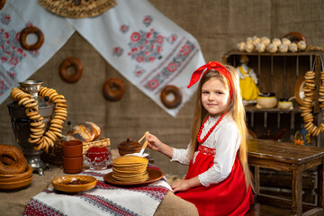 Little smiling girl in folk costume sprinklers pancakes with honey while celebrating Maslenitsa 