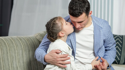Loving father shows love and embraces precious daughter at home. Lovely junior schoolgirl kisses father on cheek and hugs man tight on sofa in living room