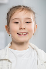 Portrait of junior schoolgirl demonstrating smile and enjoying life. Brown-haired pretty girl shows positive emotions against white shelves in kitchen