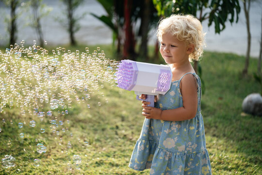 Portrait of a Child of a curly blonde girl playing with soap bubbles from a large air pistol. Outdoors in summer.