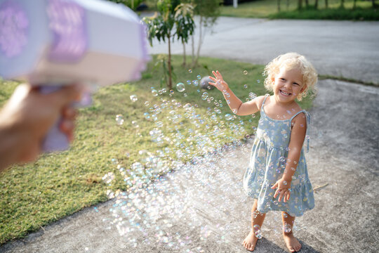 Portrait of a Child of a curly blonde girl happily playing with soap bubbles from a large air pistol with an adult. Summer outdoors under the sun.