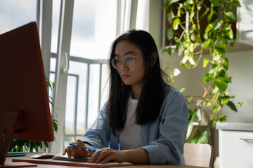 Focused Asian woman IT developer wearing glasses writing code while working remotely from home, typing on keyboard looking at desktop computer coding programming while working remotely from home