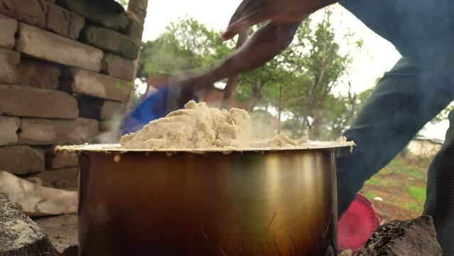 Man cooking local food ugali for students in rural school in Africa. Mixing it with blue plastic plate and wooden spoon.