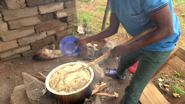 Man cooking local food ugali for students in rural school in Africa. Mixing it with blue plastic plate and wooden spoon.