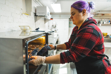 Female baked with purple hair wearing black apron putting tray of chocolate cupcakes into the oven. Professional baking. Horizontal indoor shot. High quality photo