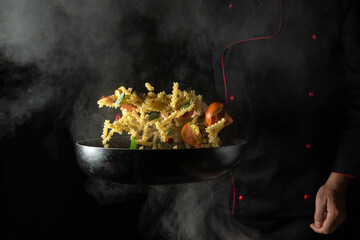 Professional chef prepares food in a hot frying pan with steam on a dark background. The concept of restaurant and hotel service. Thai food noodles with spices and vegetables