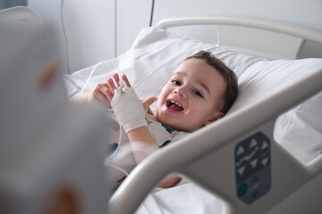 little boy in hospital lying in bed with an intravenous line in his hand smiling
