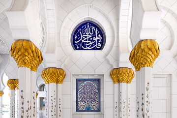 A white marbe colonnade with golden capitals leading to a quran inscription with white characters on blue background at Sheik Zayed mosque