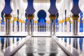 Symmetrical nightshot of the colonnade of the Sheik Zhayed mosque, with a marble catwalk surrounded by water