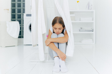 Lonely thoughtful sad small girl sits on floor in washing room near clothes dryer, wears trousers and white socks, washing machine, laundry basket and console in background, waits for parents