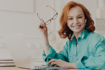 Glad red haired woman in blue shirt, works on laptop, uses earphones for watching webinar or online...