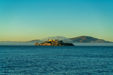 Fototapeta premium Distant alcatraz insland on the ocean with mist and fog and background mountains in golden sunset or sunrise