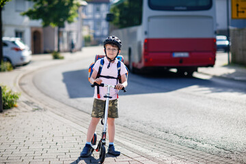 Active school kid boy with glasses in safety helmet riding with his scooter in the city with backpack on sunny day. Happy child biking on way to school. Safe way for kids outdoors to school.