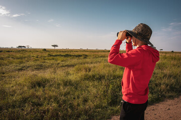 Man looking through binoculars in green savannah landscape in Serengeti National Park, Tanzania, Africa - obrazy, fototapety, plakaty