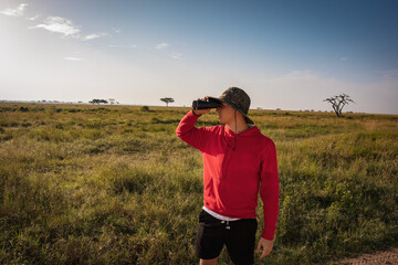 Man looking through binoculars in green savannah landscape in Serengeti National Park, Tanzania, Africa