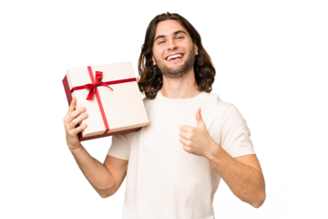 Young handsome man holding a gift over isolated background with thumbs up because something good has happened