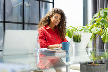 Attractive cheerful business woman in red shirt working on laptop at modern office.